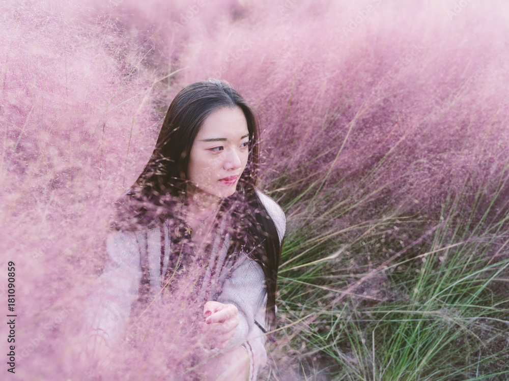 Portrait of beautiful young Chinese woman wearing white sweater sitting in the pink hairawn muhly field.