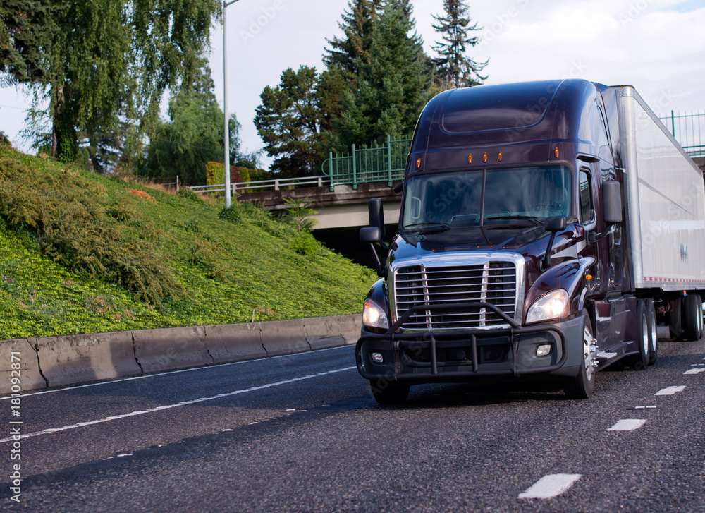 Dark modern big rig semi truck with grille guard and dry van semi trailer move with commercial cargo on straight wide highway road Dark modern big rig semi truck with grille guard and dry van semi trailer move with commercial cargo on straight wide highway road