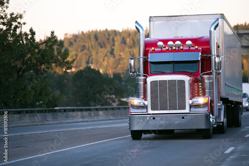 Bright red classic big rig semi truck with trailer move on evening road with turn on headlight