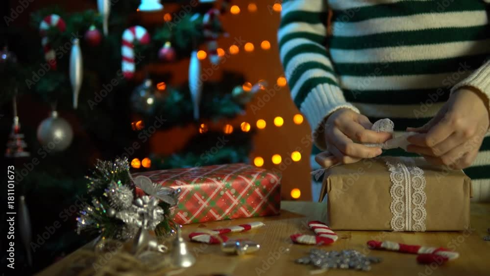 A young woman in a striped sweater tie an openwork white bow on a box with a gift next to a Christmas tree in a room decorated with yellow electric lights. A young woman in a striped sweater tie an openwork white bow on a box with a gift next to a Christmas tree in a room decorated with yellow electric lights.