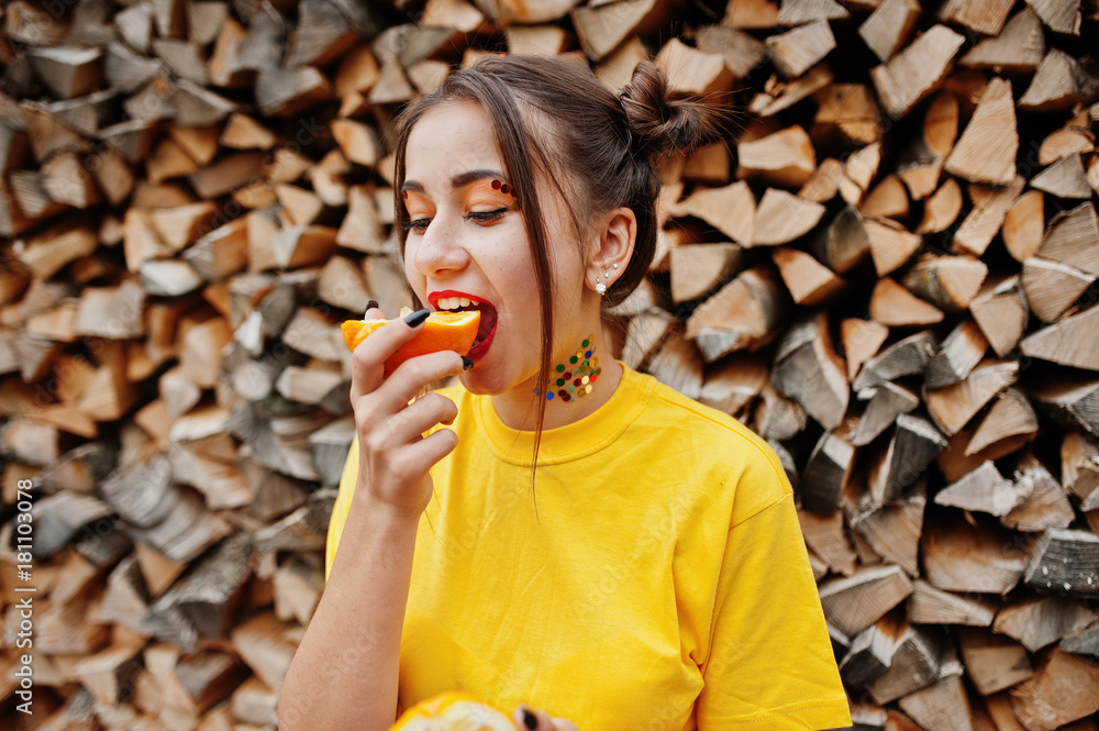 Young funny girl with bright make-up, wear on yellow shirt eat picece of orange against wooden background. Young funny girl with bright make-up, wear on yellow shirt eat picece of orange against wooden background.