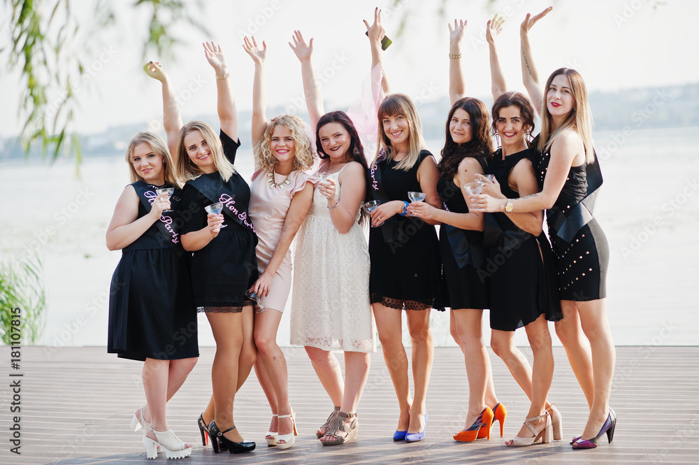 Fototapeta premium Group of 8 girls wear on black and 2 brides at hen party against sunny beach drinking champagne.