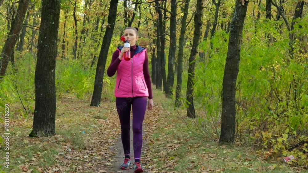 Beautiful fitness sport girl walking along the autumn park in sportswear with sports water bottle or isotonic drink in hand. Beautiful fitness sport girl walking along the autumn park in sportswear with sports water bottle or isotonic drink in hand.