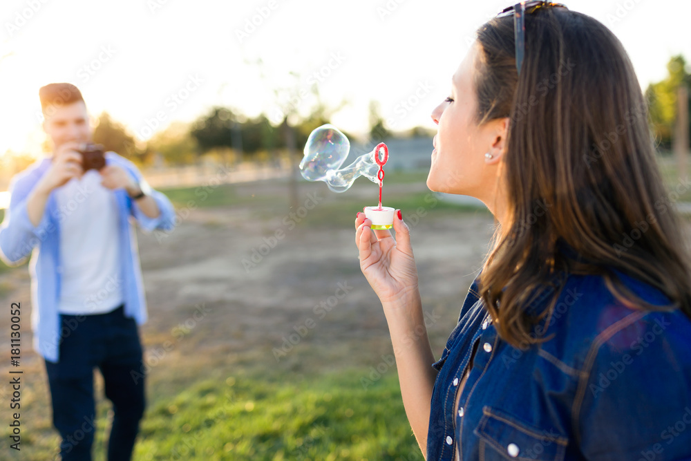 Fototapeta premium Handsome young man taking photo of his girlfriend making soap bubbles in the park.