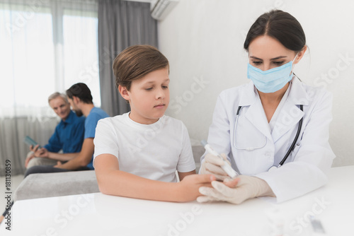 A nurse takes a blood test with a boy s scarifier. A nurse is sitting in a mask and gloves at the table.