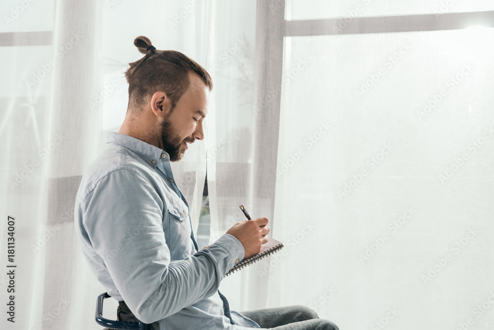 man writing in notebook in front of window