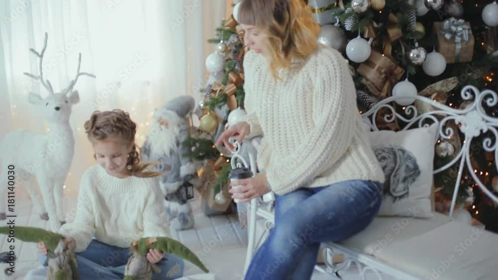 family Christmas photo session. Mom and daughter are sitting near the Christmas tree, in white knitted sweaters and jeans family Christmas photo session. Mom and daughter are sitting near the Christmas tree, in white knitted sweaters and jeans