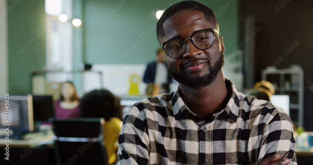 Portrait shot of young African American man in glasses turning his head to the camera and smiling in the modern office. Working people on the blurred background. Close up Portrait shot of young African American man in glasses turning his head to the camera and smiling in the modern office. Working people on the blurred background. Close up