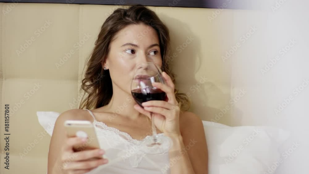 Pretty woman lies on bed with smartphone and drinks red wine out of glass Pretty woman lies on bed with smartphone and drinks red wine out of glass