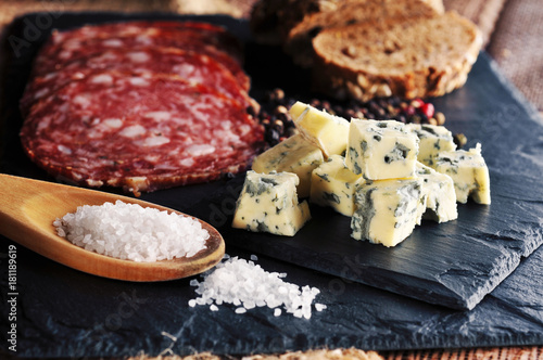 Roquefort slices with bread, sliced salami, salt on the spoon and peppercorns on the dark background