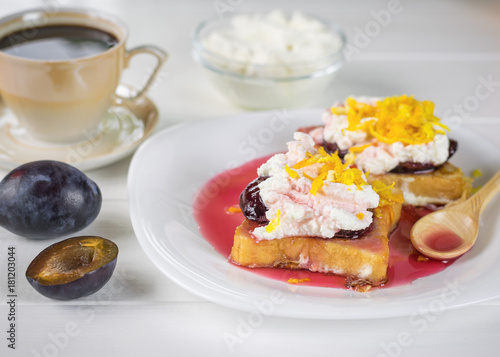 Breakfast of French bread with curd cream, plum jam and orange zest on a white table.