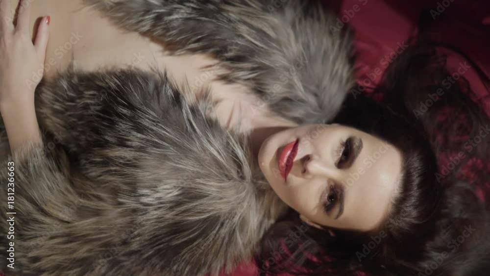 Beautiful woman in fur lying on the bed. Young brunette woman with perfect body wearing a fur coat lies on her back across the red bed sheets. Woman looking at camera. View from above. Beautiful woman in fur lying on the bed. Young brunette woman with perfect body wearing a fur coat lies on her back across the red bed sheets. Woman looking at camera. View from above.