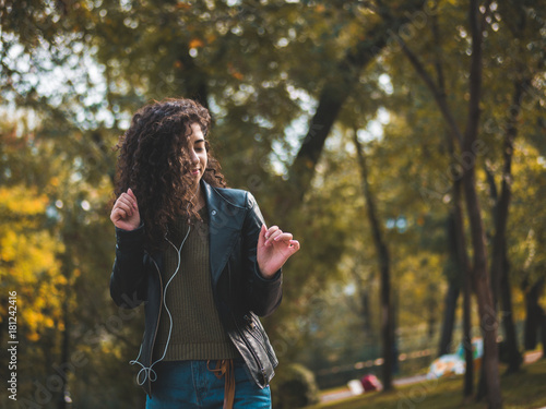 Happy african young woman with curly hair listening to music on earphones. Hispanic hipster girl dancing to rhythm and singing along melody in the autumn park.