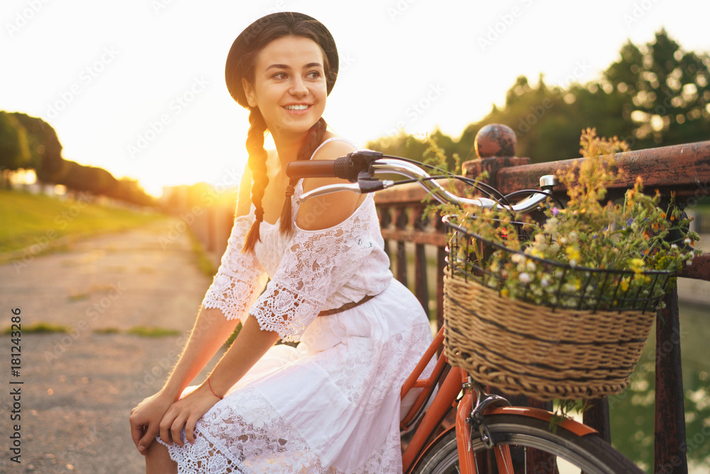 Fototapeta premium Young beautiful woman sitting on her bicycle with flowers at sunset