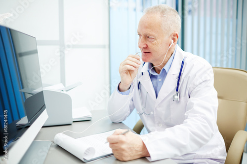 Contemporary doctor in earphones consulting patients through video calling while sitting in front of computer monitor by workplace