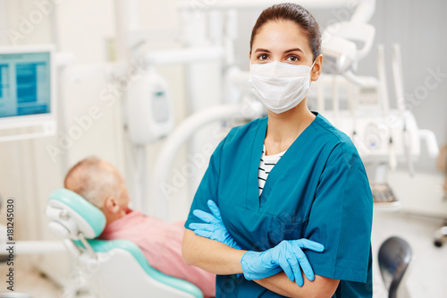 Modern orthodontist in uniform  mask and gloves looking at camera with her patient waiting on background