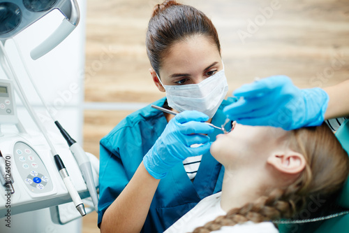 Young dentist in uniform examining teeth of little girl with medical tools