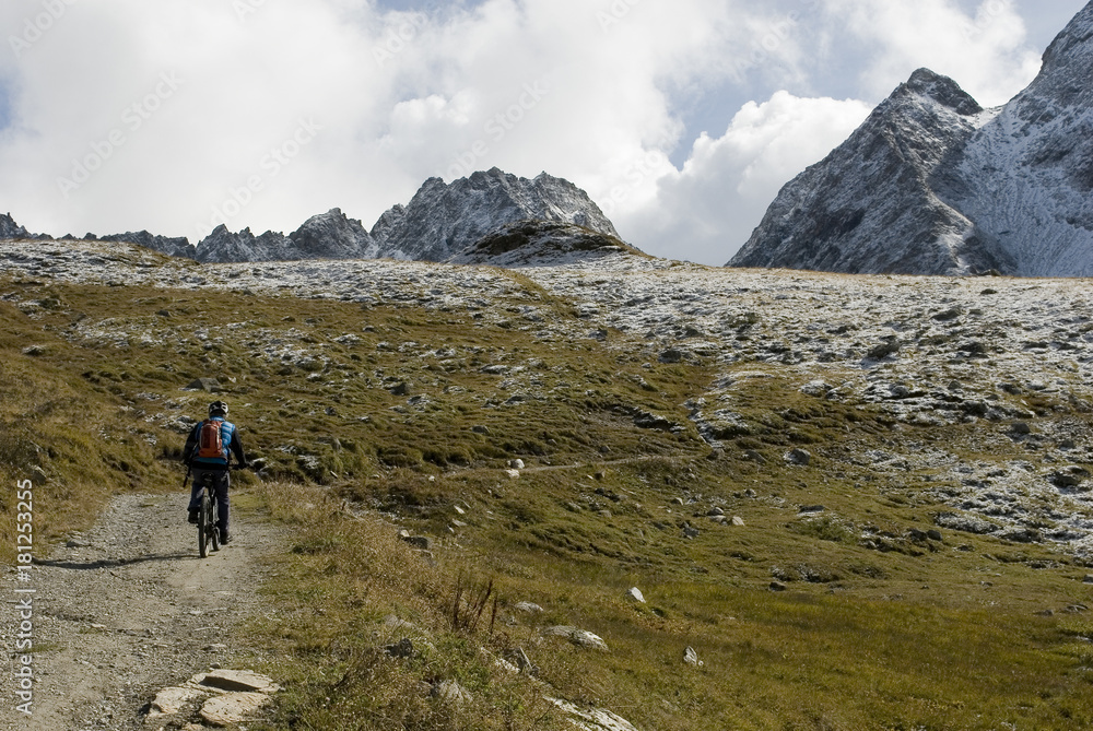 Fototapeta premium man with an ebike, electric bike, mountains covered with fresh snow just fall, autumn, observes Kastelhorn (Castel Peak), grass, dirt, October, Alps, light, clouds, Formazza valley, Piedmont, Italy