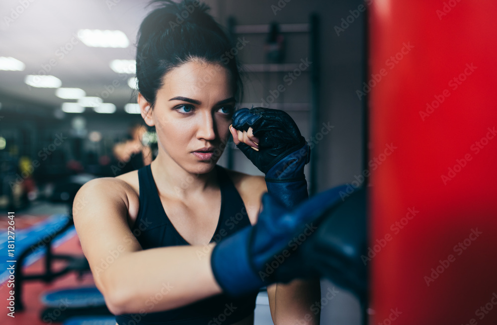 Caucasian beautiful brunette sportswoman trainer punching a red bag with kickboxing gloves in the gym. Sport, fitness, lifestyle and motivation concept.