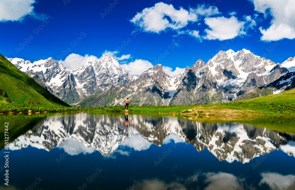 Fototapeta premium Mirror Lake on the background of snowy mountains. Blue sky with white clouds. Human silhouette and mirror image of nature in the lake. Summer. Georgia. Svaneti, Mestia
