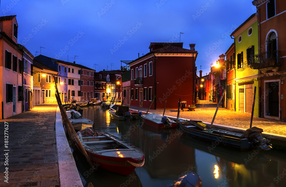 Quiet evening cityscape of Burano island in Venice. Rainbow colors of small cottages. Dark blue sky. Small boats are parked along the water canal.