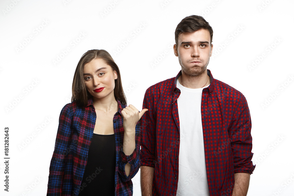 Portrait of charming young lady wearing red lipstick smiling and pointing finger at her irritated bearded man who is blowing cheeks, expressing displeasure, irritation, dislike and annoyance Portrait of charming young lady wearing red lipstick smiling and pointing finger at her irritated bearded man who is blowing cheeks, expressing displeasure, irritation, dislike and annoyance