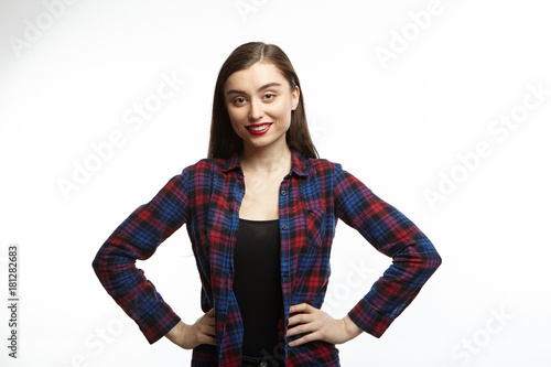 Studio shot of attractive self confident young female in great mood feeling happy, holding hands on her slender waist and looking at camera with radiant smile, rejoicing at good day or news