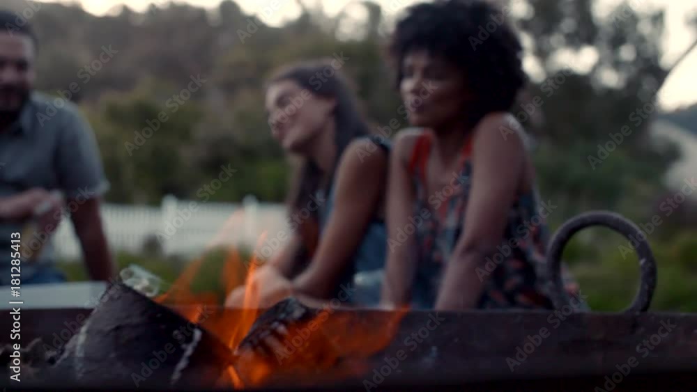 Group of young men and women sitting together in backyard near campfire with drinks. Young friends having a party outdoors. Group of young men and women sitting together in backyard near campfire with drinks. Young friends having a party outdoors.