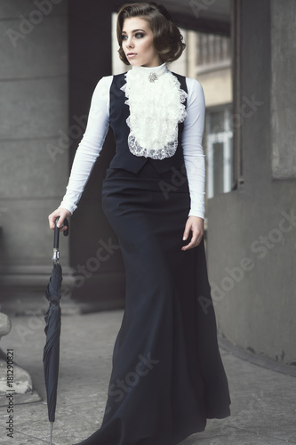 Portrait of gorgeous woman with elegant Victorian hairstyle wearing old-fashioned gown with jabot walking among the pillars of her old castle leaning on black umbrella. Vintage style. Outdoor shot