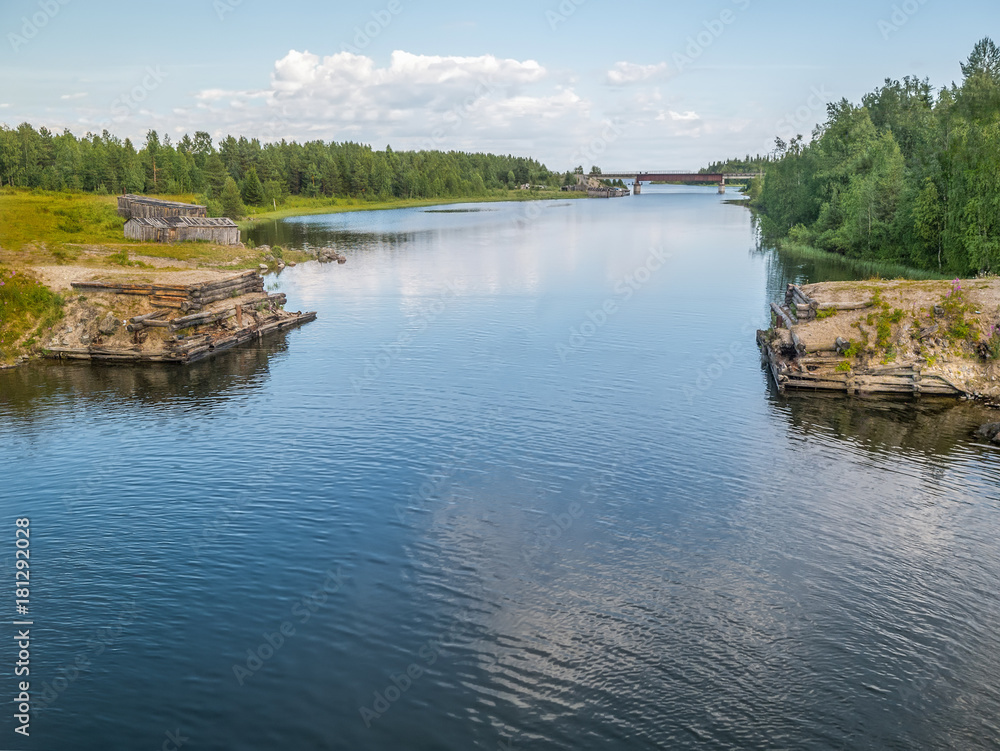 Fototapeta premium Summer water landscape. Blue sky and clear water of river Sofyanga with reflected clouds. Old wooden authentic buildings on the river bank