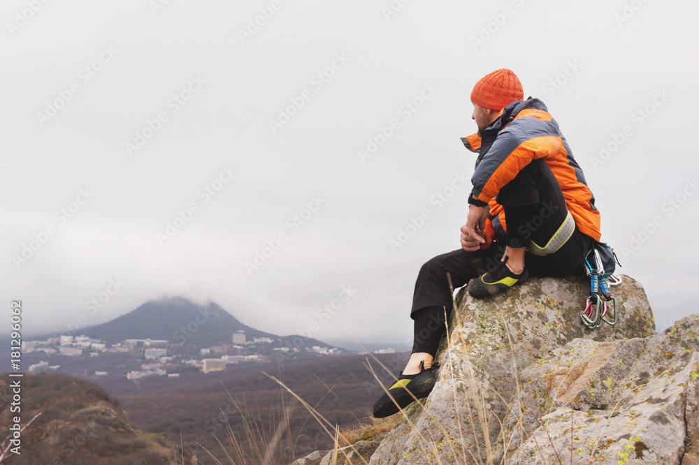 Obraz premium Hipster - a climber in a down jacket and a knitted cap sits and rests on the top of a rock