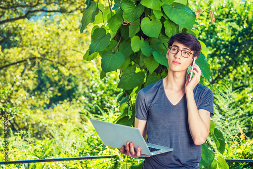 Growing with Nature. Asian American college student studying in New York. 20 years old man stands by green big leaf plants on campus, works on laptop computer, talks on cell phone. Way to Success..