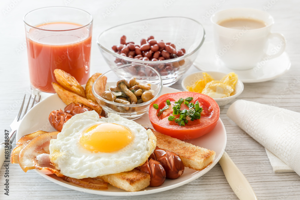 Full English breakfast with smoked sausages, fried egg, bacon, tomato, toast and beans. Tea with milk. A glass of fresh juice. Cutlery. Breakfast on a white plate on a light background. soft light