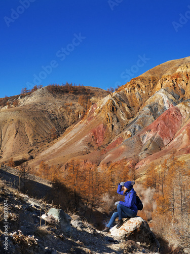 A young tourist is sitting on a rock and looking at the beautiful mountain topography. Multicolored paint of rock. A hiking woman with a backpack relaxing atop a rock enjoying a sunny autumn afternoon