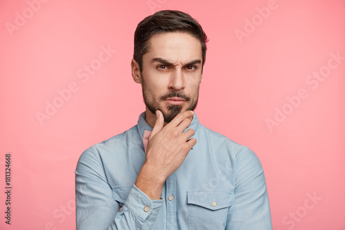 Headshot of serious pensive unshaven pleasant looking male businessman keeps hand on chin, has clever look, tries to understand details of contract, frowns face, looks confidently and strict