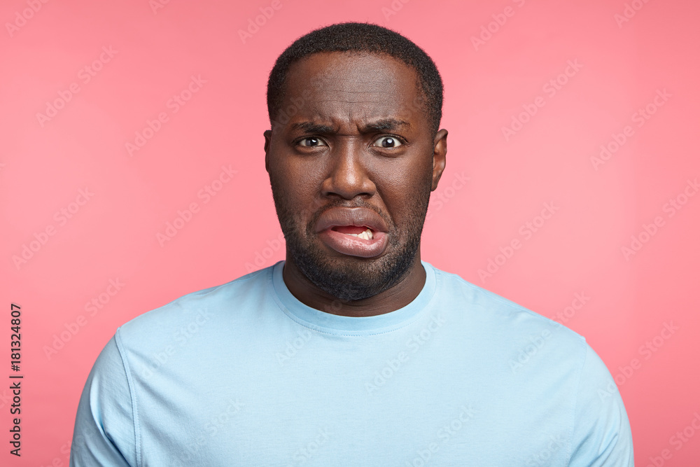 Discontent young African American male curves lips, dislikes what he sees, displeased with bad news, has disgust towards something, isolated over pink background. Negative human emotions concept. Discontent young African American male curves lips, dislikes what he sees, displeased with bad news, has disgust towards something, isolated over pink background. Negative human emotions concept.