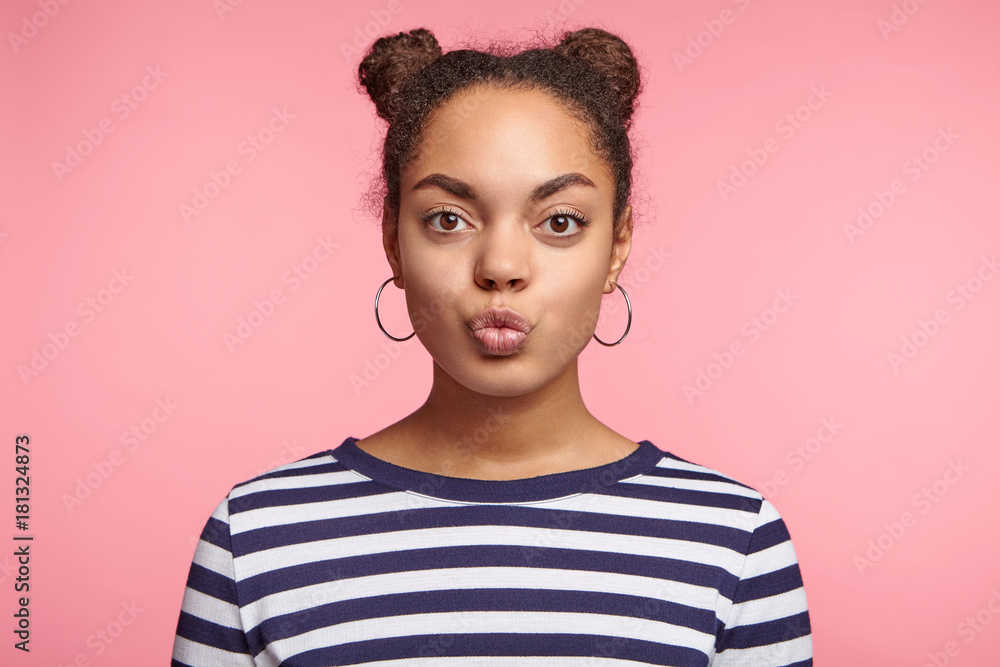 Cute female teenager with black skin, two hair buns, wears earrings and srtiped sweater, rounds lips, looks coquette, isolated over pink background. Fashionable stylish female going to recieve kiss Cute female teenager with black skin, two hair buns, wears earrings and srtiped sweater, rounds lips, looks coquette, isolated over pink background. Fashionable stylish female going to recieve kiss