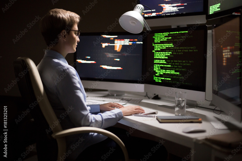 Profile view of confident middle-aged programmer in eyeglasses sitting in front of modern computer and writing programming code, interior of dim office on background