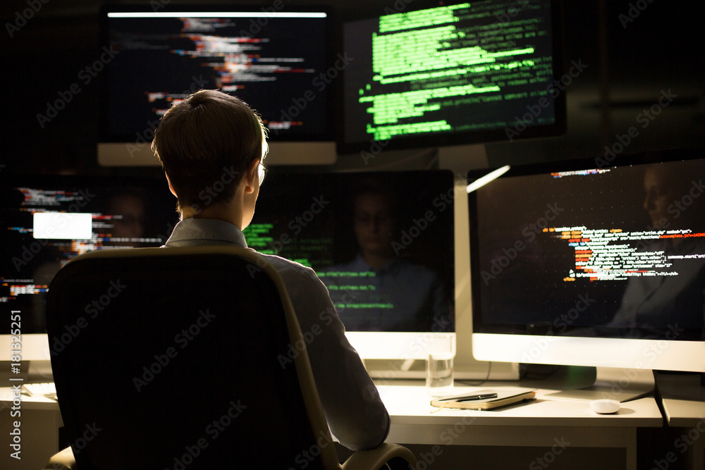 Working process in dim office: female programmer wearing white shirt wrapped up in coding while sitting at desk, rear view Working process in dim office: female programmer wearing white shirt wrapped up in coding while sitting at desk, rear view