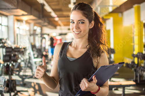 Smiling female fitness instructor with clipboard showing thumb up in gym.