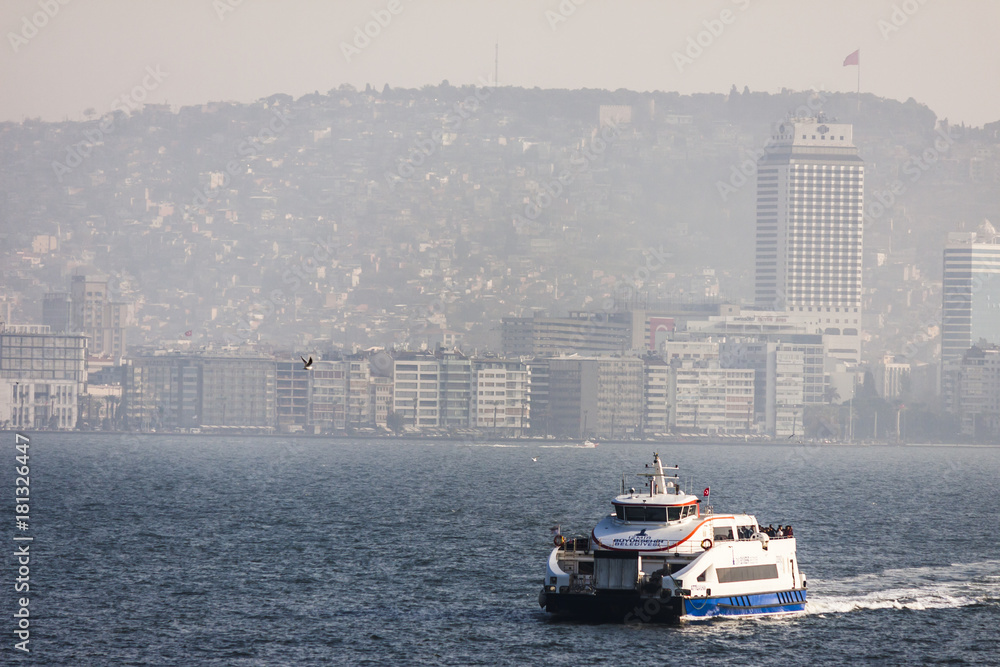 A ferry boat is navigating on the sea in front of Izmir: the panorama of the city climbing the hills in a soft white fog of an autumn sunny day. Izmir (Turkey); 16 november 2017