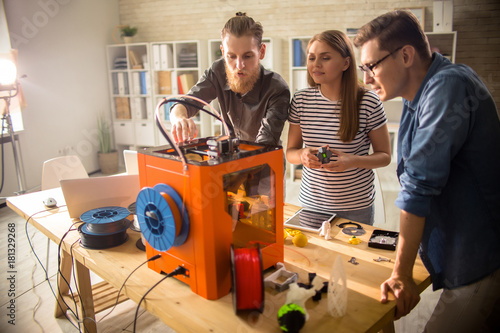Three young engineers standing at wooden desk and using 3D printer in order make small prototypes, interior of modern laboratory on background