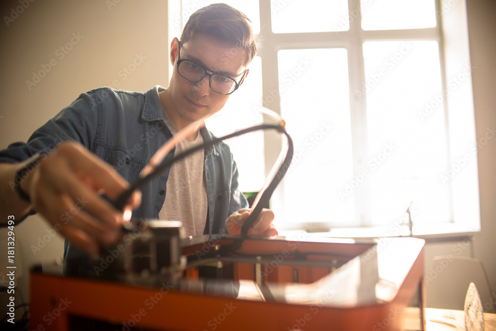 Waist-up portrait of concentrated young technician in eyeglasses repairing 3D printer after breakage, interior of spacious laboratory on background Waist-up portrait of concentrated young technician in eyeglasses repairing 3D printer after breakage, interior of spacious laboratory on background