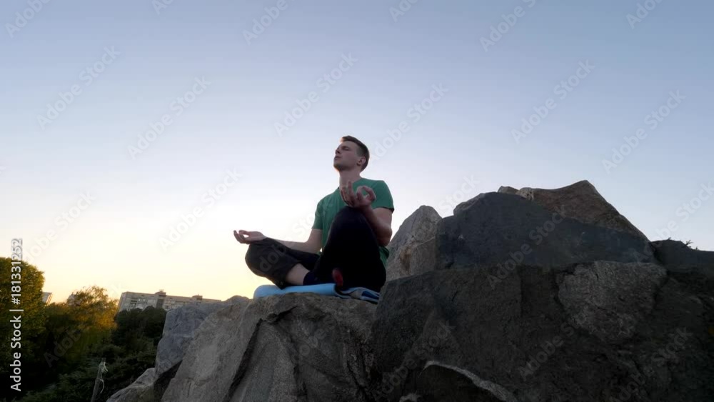 Young man sits on top of a hill meditating at sunset in a park, tilt shot Young man sits on top of a hill meditating at sunset in a park, tilt shot
