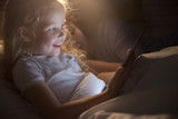 Portrait of adorable little girl smiling happily  using digital tablet lying in bed at night, face lit by screen light like magic, copy space