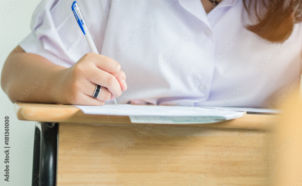 Fototapeta premium Girl Students holding pen taking exams, writing examination room with undergraduate students inside. Student sitting learning lessons on row desk doing final exam in classroom with uniform.
