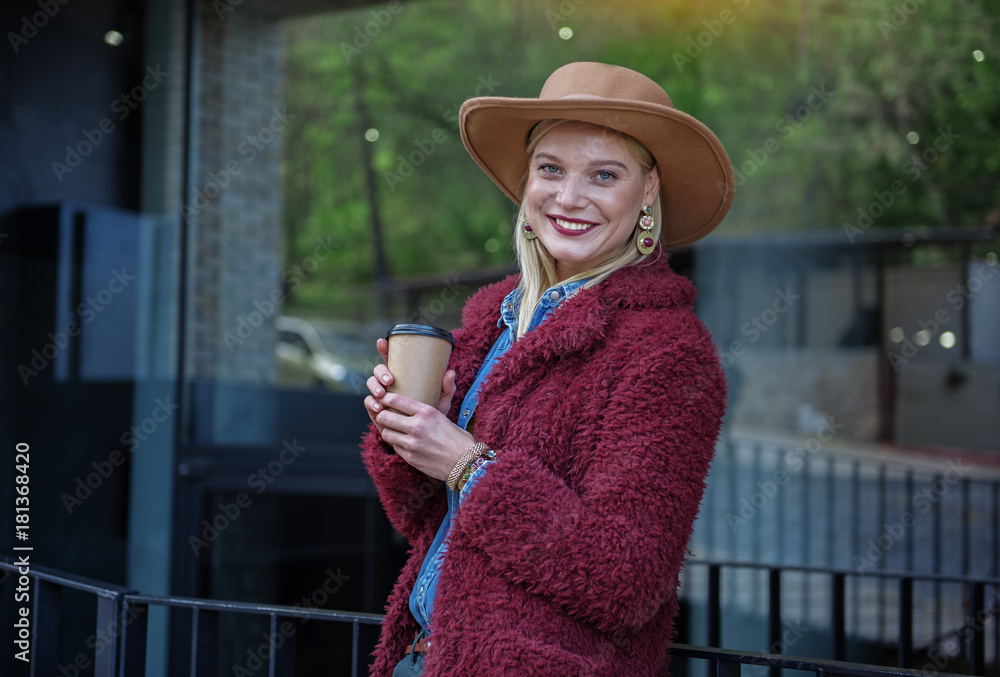 Waist up portrait of happy blond girl drinking coffee in city outdoor. She is leaning on border with relaxation and laughing. Copy space