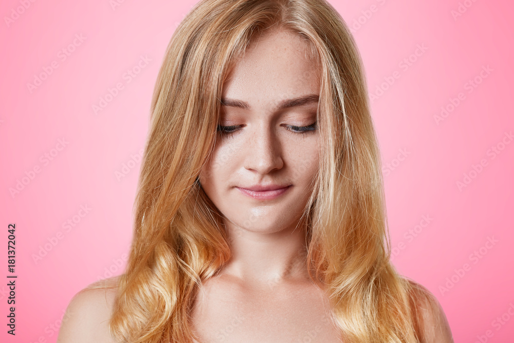 Naklejka premium Serious blonde female with appealing apearance looks down, has shy expression, isolated over pink background. Good looking young woman has healthy pure skin models in studio. Skincare concept