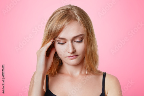 Serious pleasant looking young female model keeps hand on temples, looks pensively down, has thoughtful expression, tries to remember something, has headache after long working day, rests at home