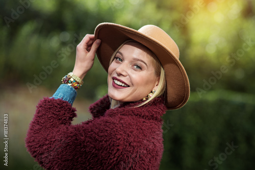 Portrait of happy blond girl is smiling while looking at camera with joy. She is standing outdoor while touching hat elegantly